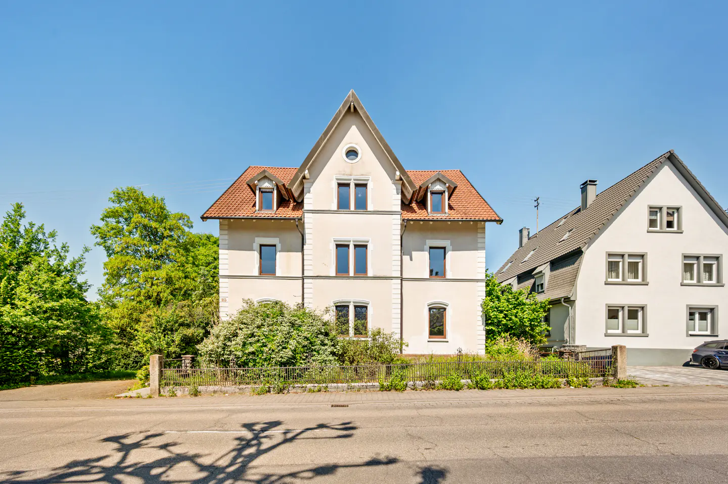 Two-story beige house with a red tile roof, dormers, and a pointed gable, viewed from across the street on a sunny day.