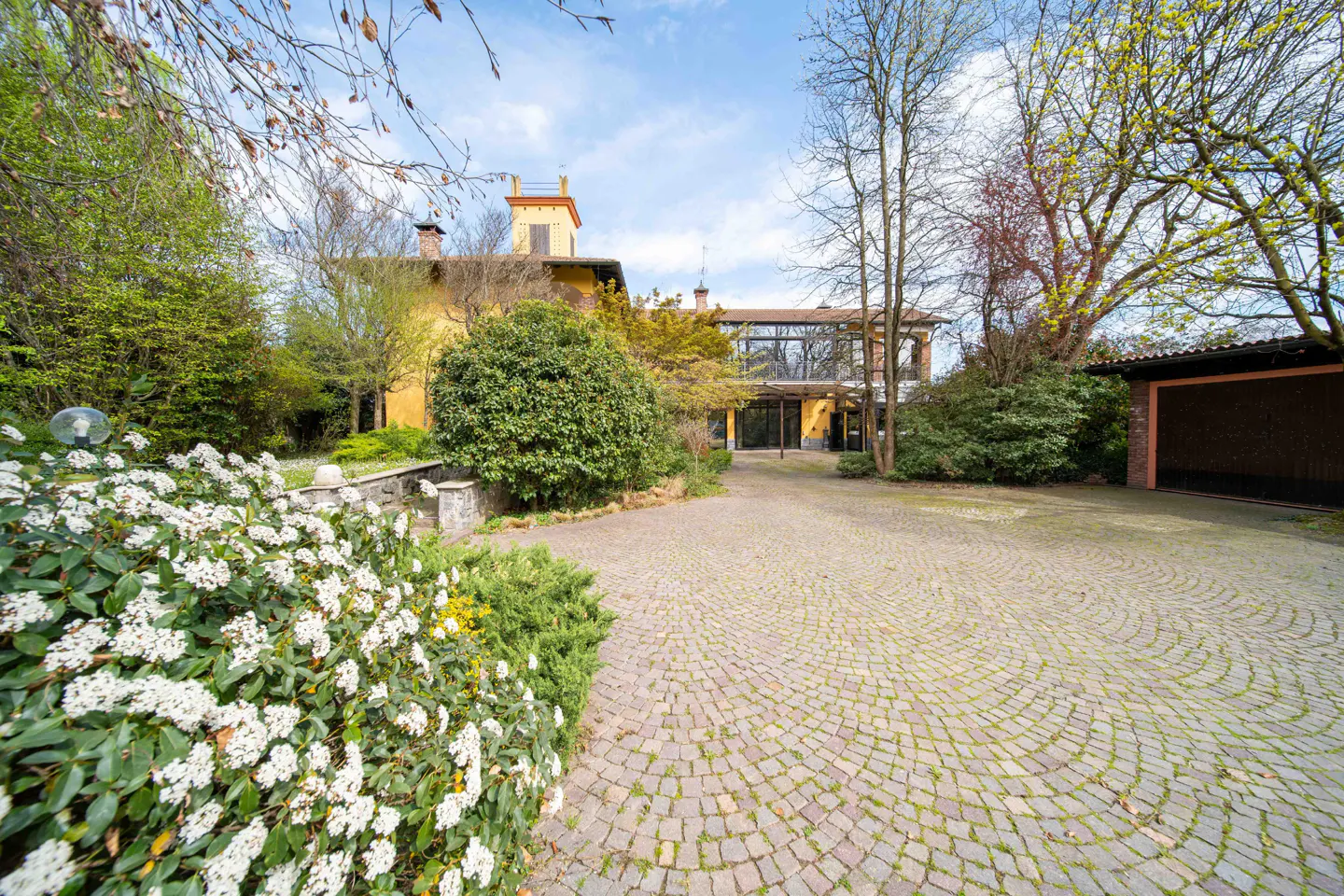 Exterior view of a yellow house with a tower, surrounded by trees and a circular brick driveway. White flowers in the foreground.