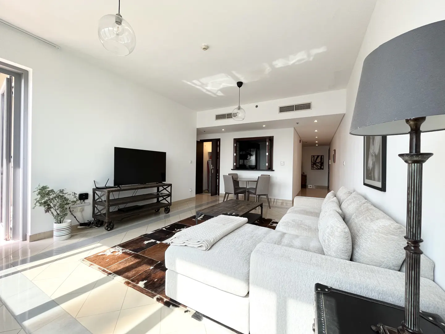 Bright living room with white walls, a large gray sofa, a TV on a metal stand, and a dining area in the background.