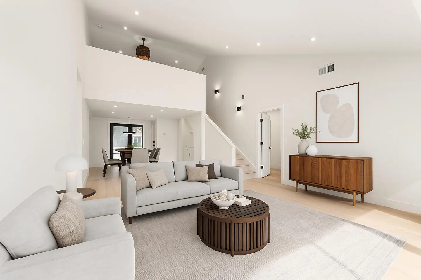 Bright, modern living room with white walls, light wood floors, and gray sofas. A round wood coffee table sits on a light gray rug. Dining area visible in the background.