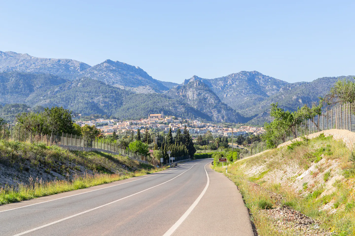 Asphalt road leading to a town nestled in front of mountains under a clear blue sky.