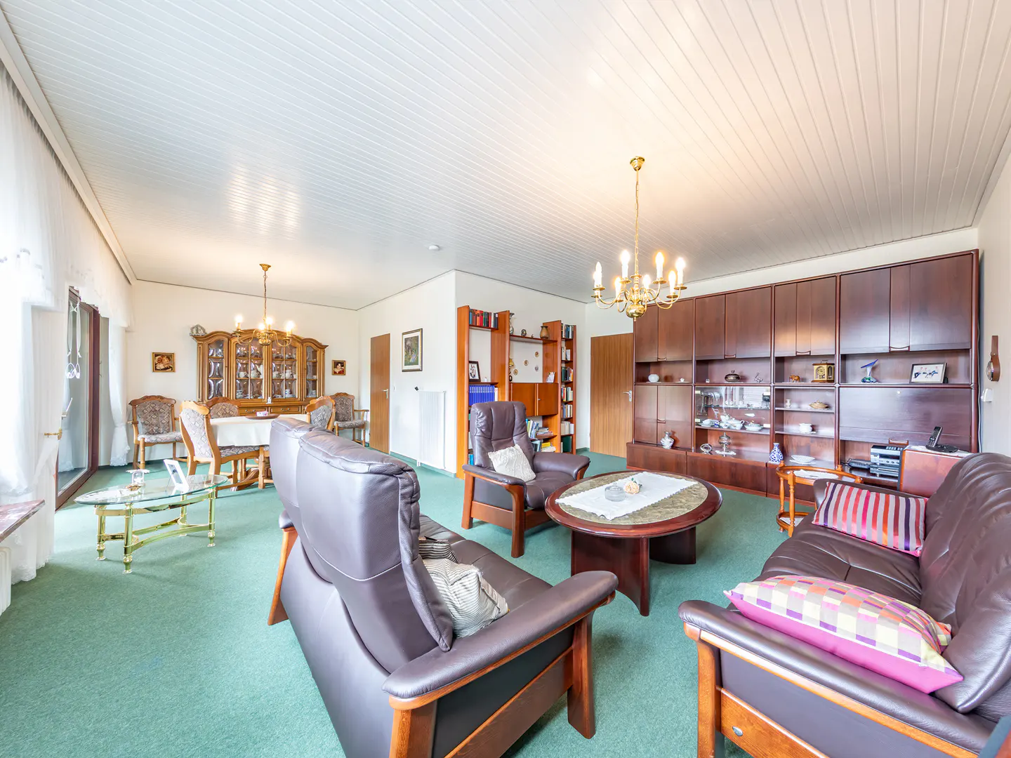 A wide shot of a living room with brown leather furniture, a green carpet, and a dining area in the background.