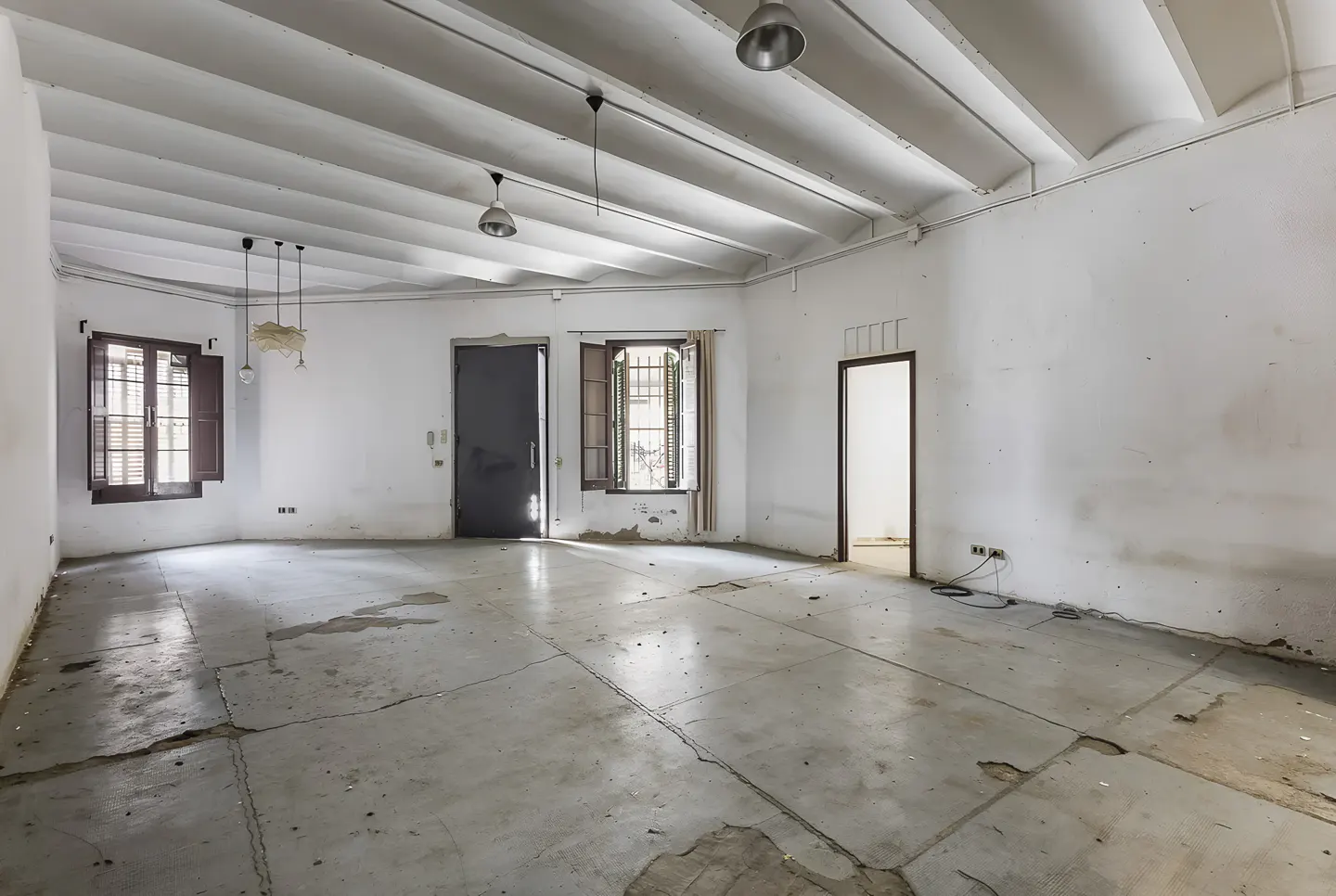 An empty room with white walls, a concrete floor, and two windows with brown shutters. There are three lights hanging from the ceiling.