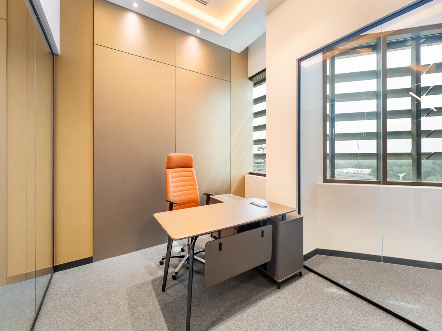 A modern office space with a tan desk, orange leather chair, and large windows with blinds. The walls are paneled in neutral tones.