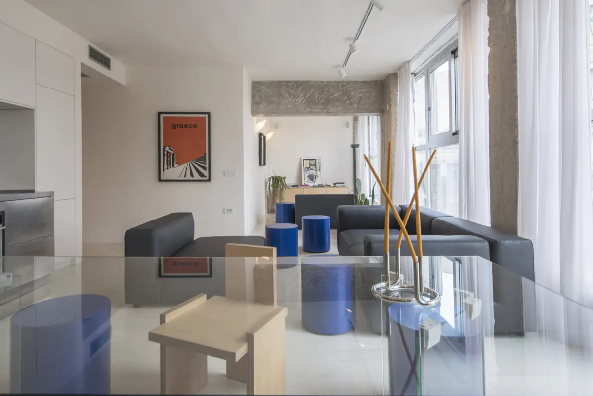 Bright, modern living room with black sofas, blue stools, and a glass table. A "Greece" poster hangs on the white wall. Natural light streams through sheer curtains.