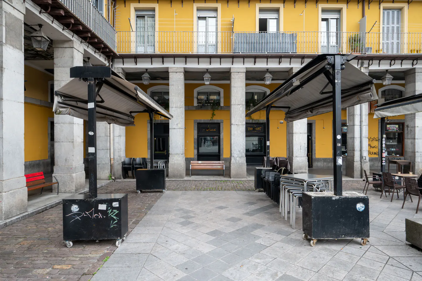 A courtyard with stone pillars, yellow walls, and outdoor cafe seating under retractable awnings on a paved plaza.