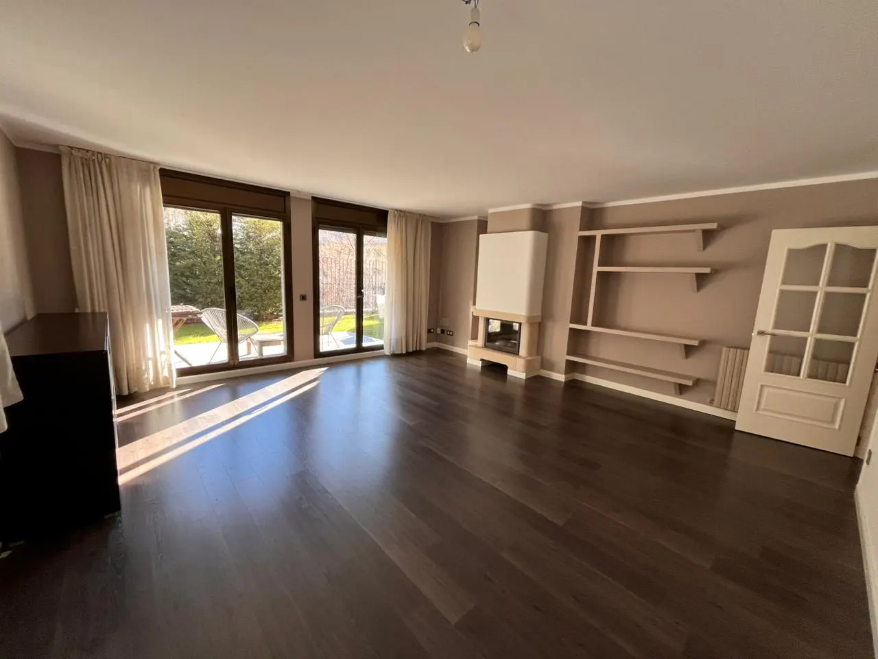 A bright, empty living room with dark wood floors, a fireplace, and large windows overlooking a green yard.