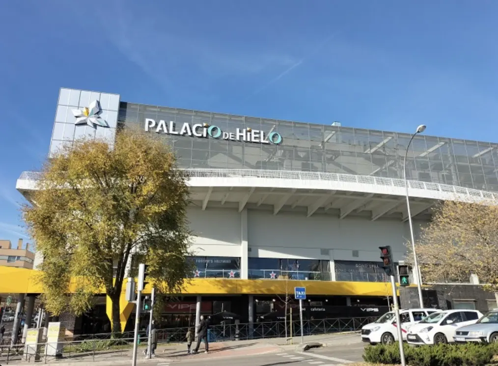 Exterior view of Palacio de Hielo, a large, modern building with "Palacio de Hielo" signage. Cars parked nearby, and a tree is on the left.