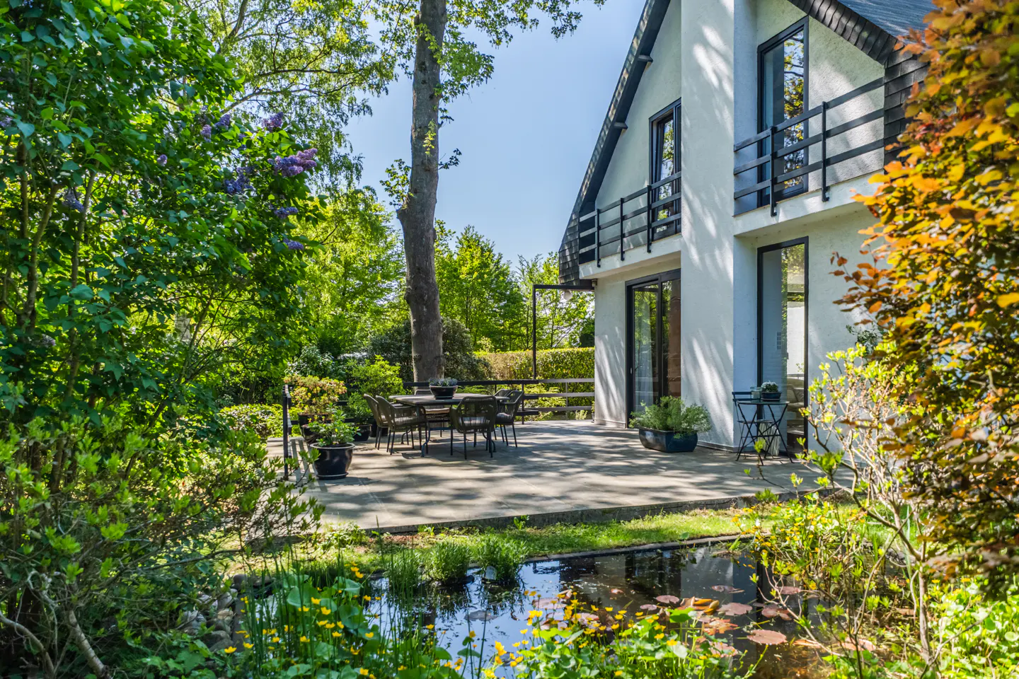 Exterior view of a white house with a patio, table, chairs, and a pond with lily pads.