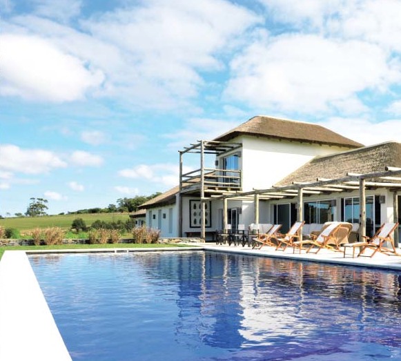 Luxury home with a thatched roof and a blue swimming pool in the foreground on a sunny day. [AI-generated text]