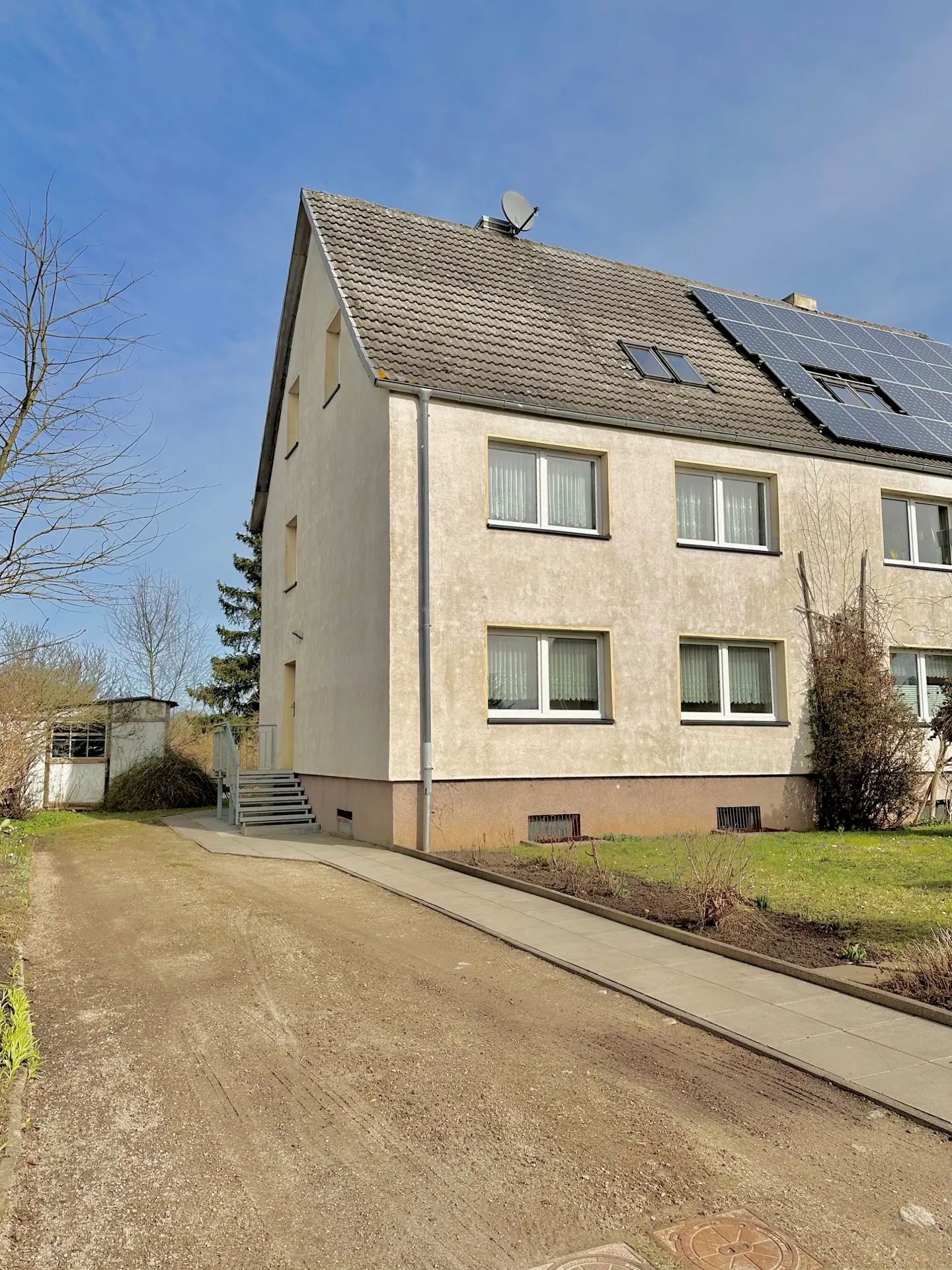 Two-story beige apartment building with a gray roof, solar panels, and a satellite dish under a blue sky.
