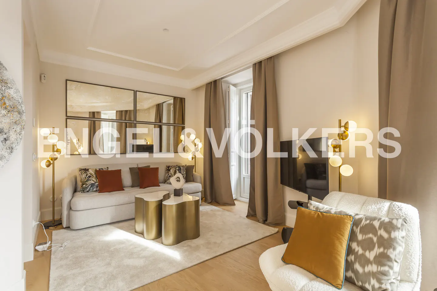 Modern living room with a beige sofa, gold coffee tables, patterned pillows, and floor lamps on a light rug.