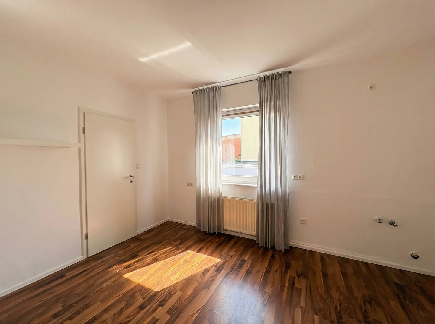 Bright, empty room with wood floors, white walls, and a window with sheer gray curtains. A white door is on the left.
