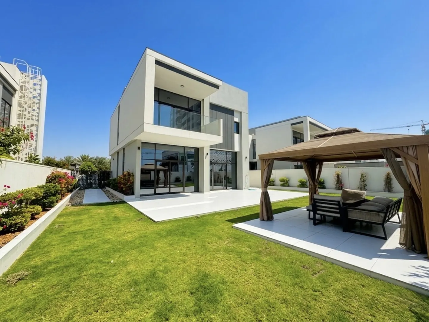 Two-story modern house with a white patio, green lawn, and a gazebo with seating under a clear blue sky.