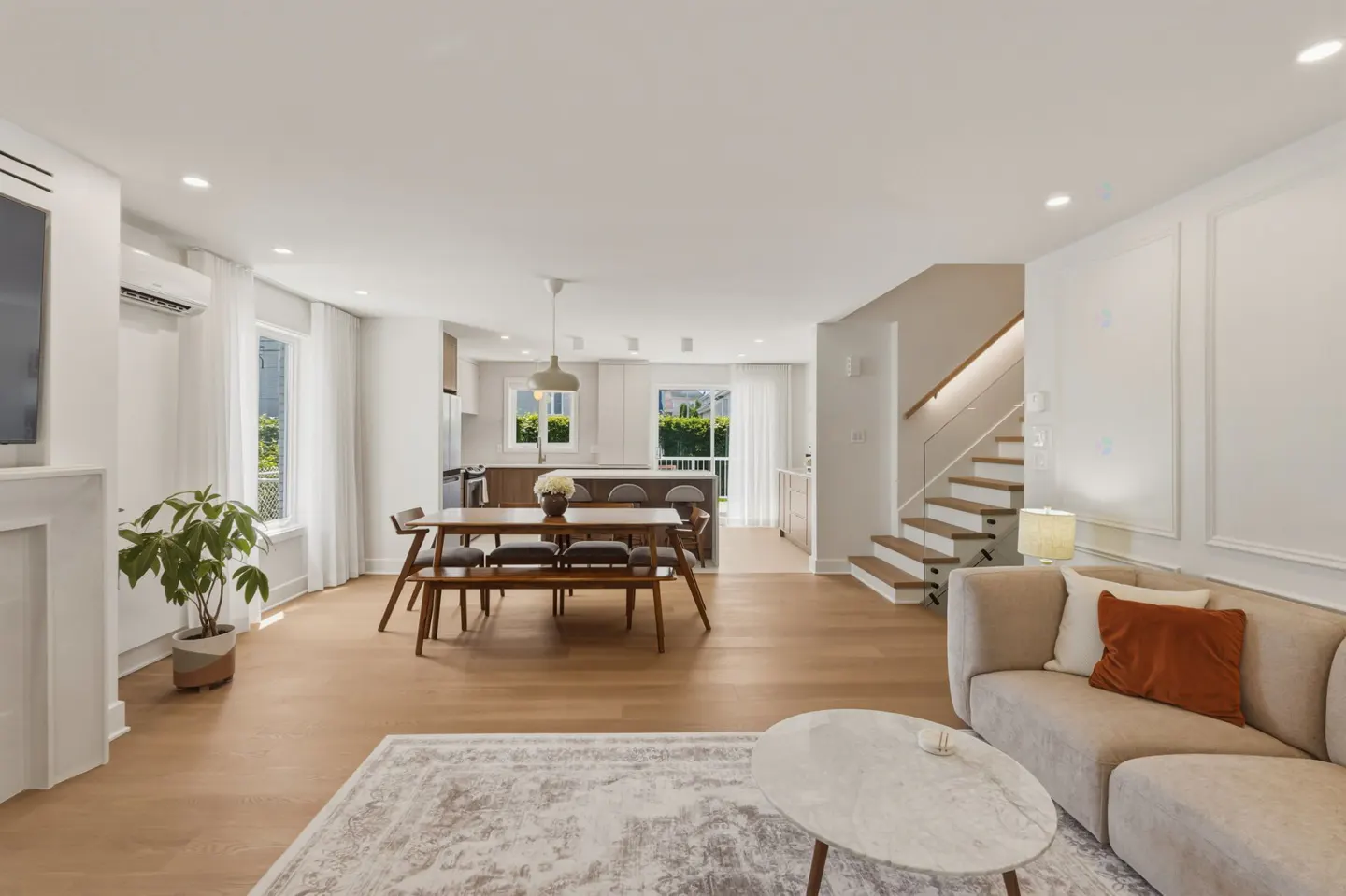 Open-concept living area with a beige sofa, marble coffee table, wooden dining set, and stairs leading to an upper floor.
