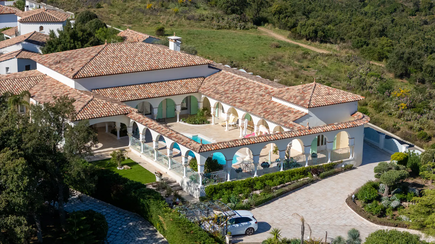 Aerial view of a white villa with a red tile roof, arched walkways surrounding a pool, and a stone driveway with a white car.