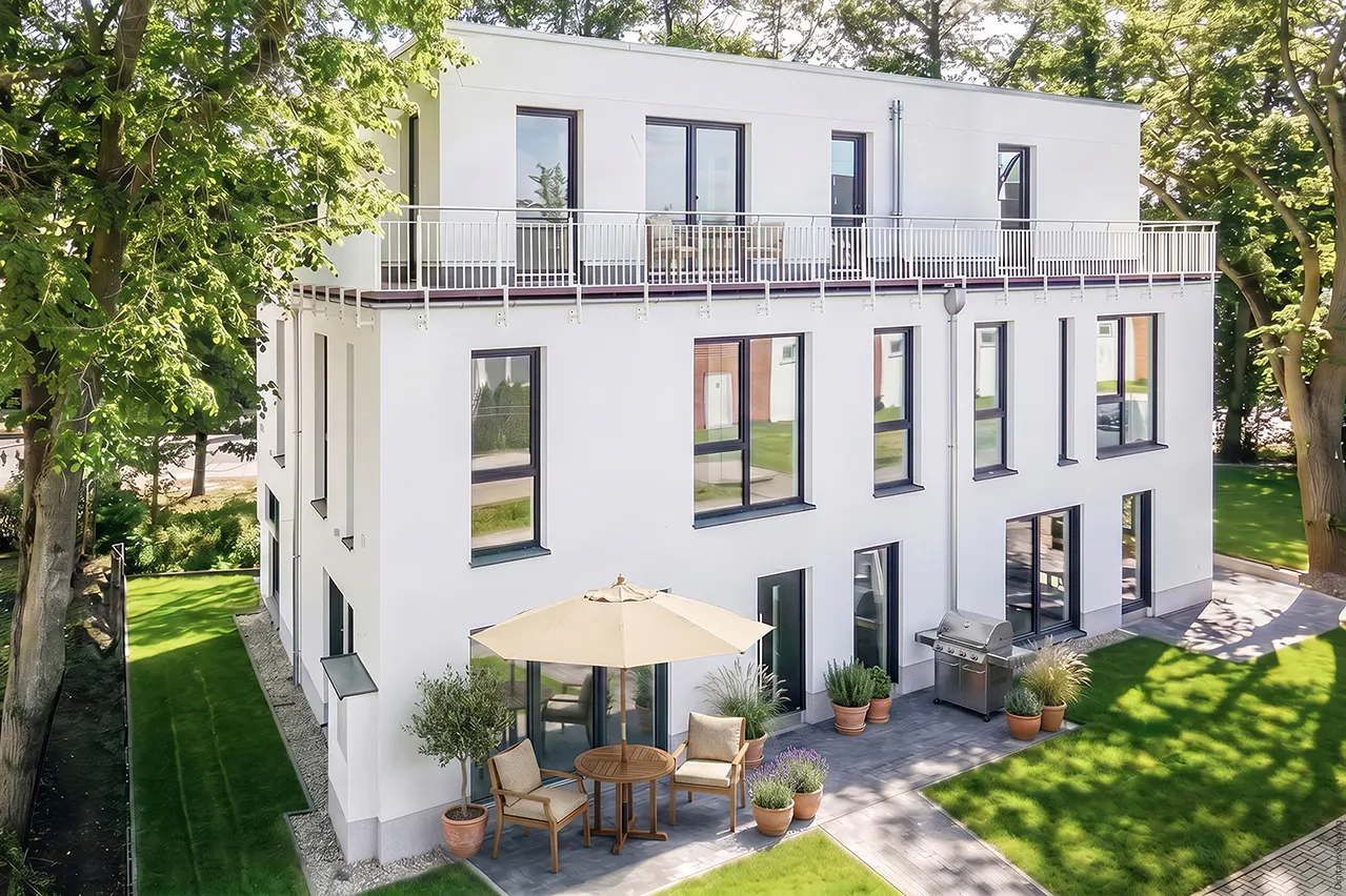 Modern white three-story house with a balcony, patio furniture, grill, and green lawn.