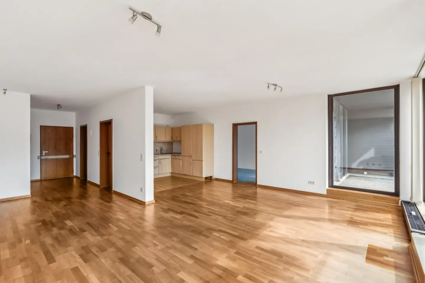 Bright, empty apartment with wood floors, white walls, and a kitchen in the background. A large window is on the right.