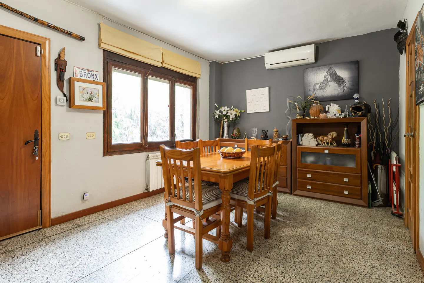 A dining room with a wooden table, chairs, and a cabinet against a gray wall. A window with a yellow blind lets in natural light.