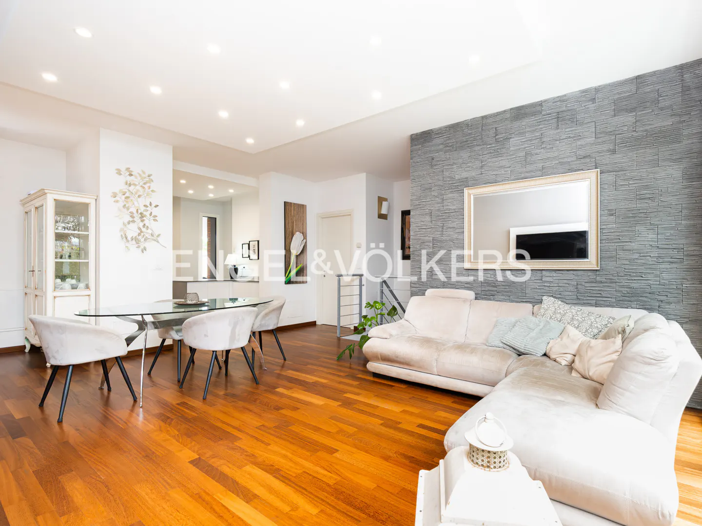 Open-concept living and dining area with wood floors, a gray stone accent wall, and a beige sectional sofa.
