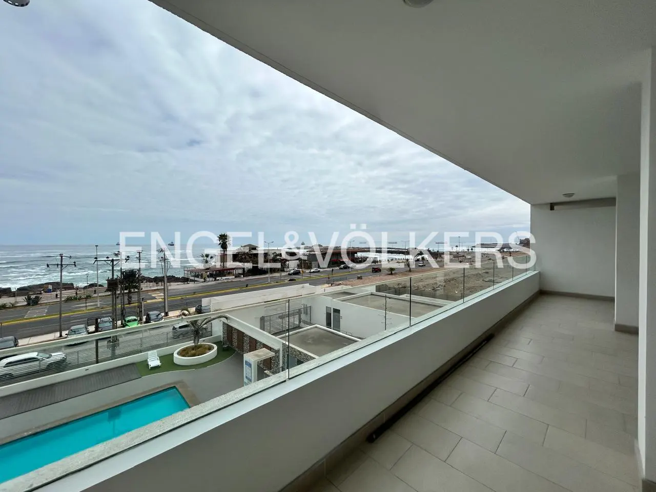 Balcony view of ocean, road, and pool. Glass railing, white walls, and tile floor. Cloudy sky. Engel & Völkers logo.
