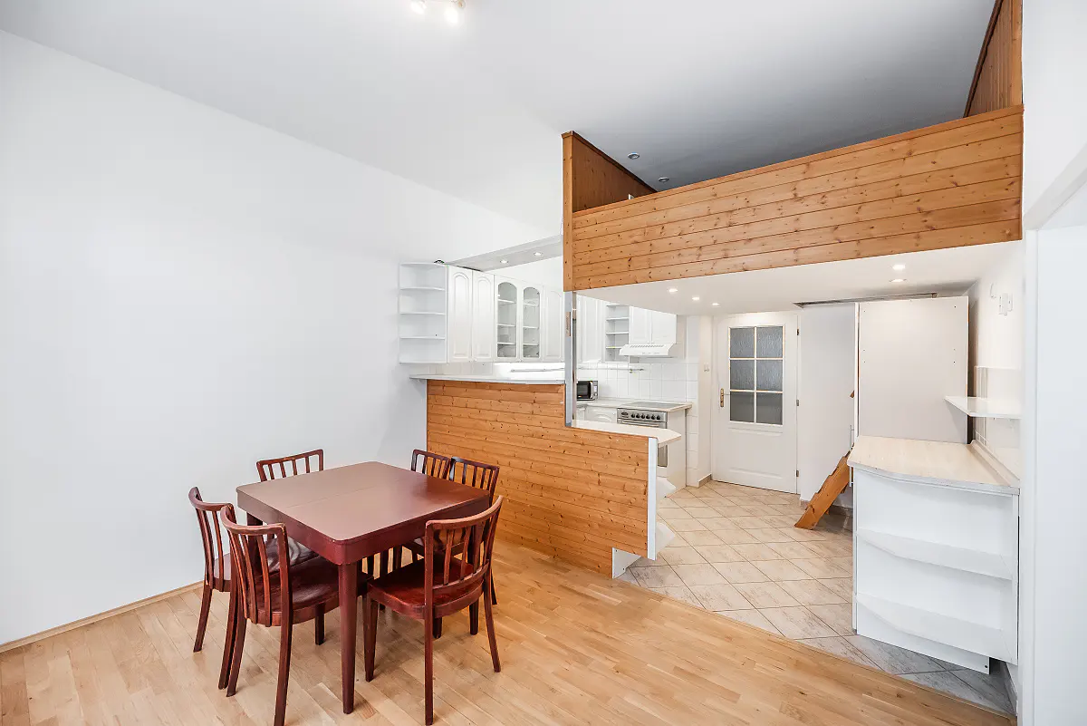 Bright apartment interior with a dining table, kitchen, and wooden loft. The walls are white, and the floor is light wood.