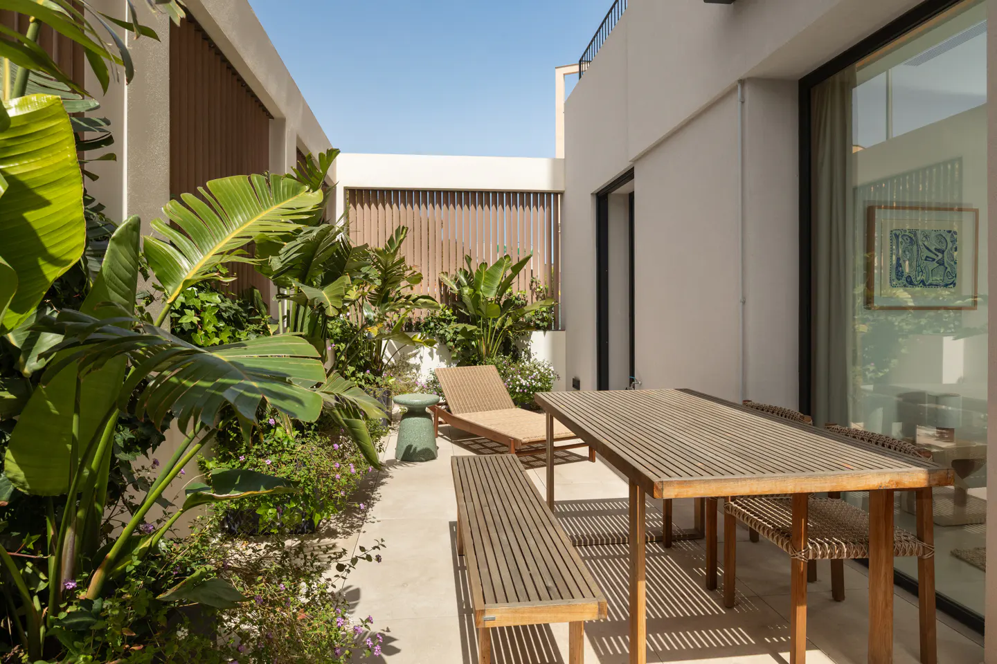 Outdoor patio with a wooden table, bench, and lounge chair surrounded by lush green plants and a white wall.