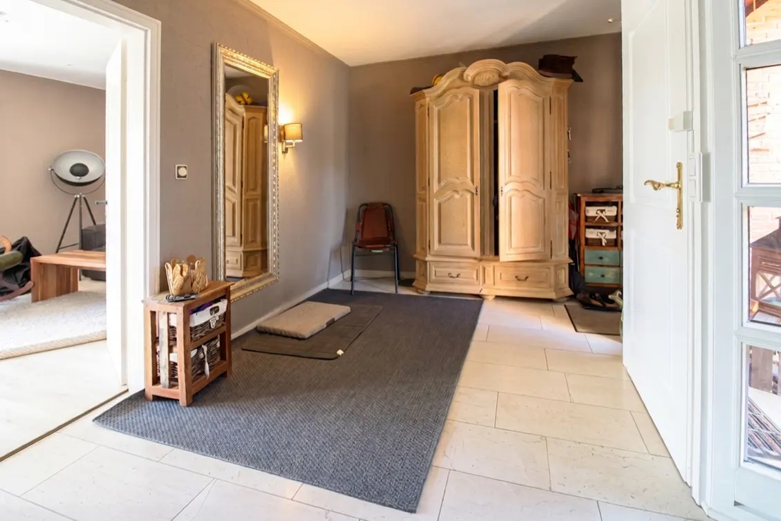 Entryway with light tile floors, a large grey rug, a wooden wardrobe, a mirror, and a chair, leading to an adjacent room.