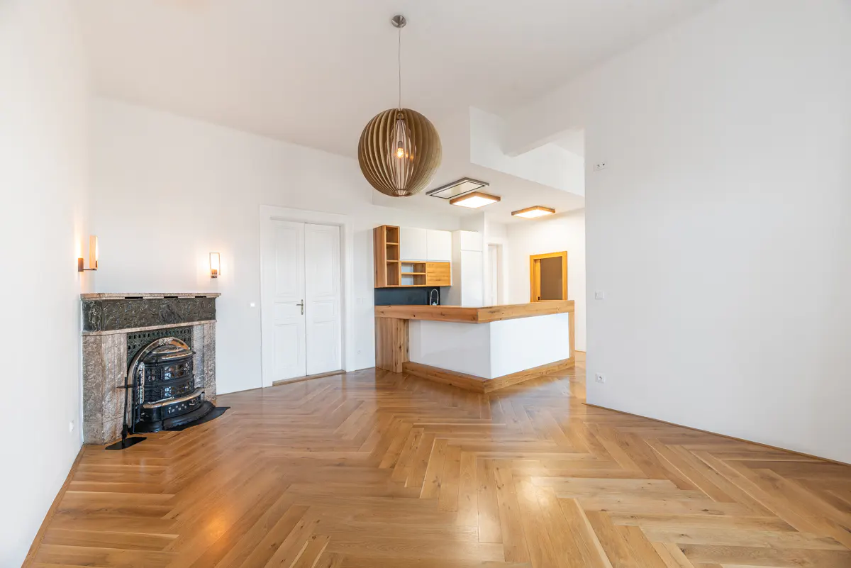 Bright, empty room with herringbone wood floors, white walls, and a fireplace. A kitchen area is visible in the background.