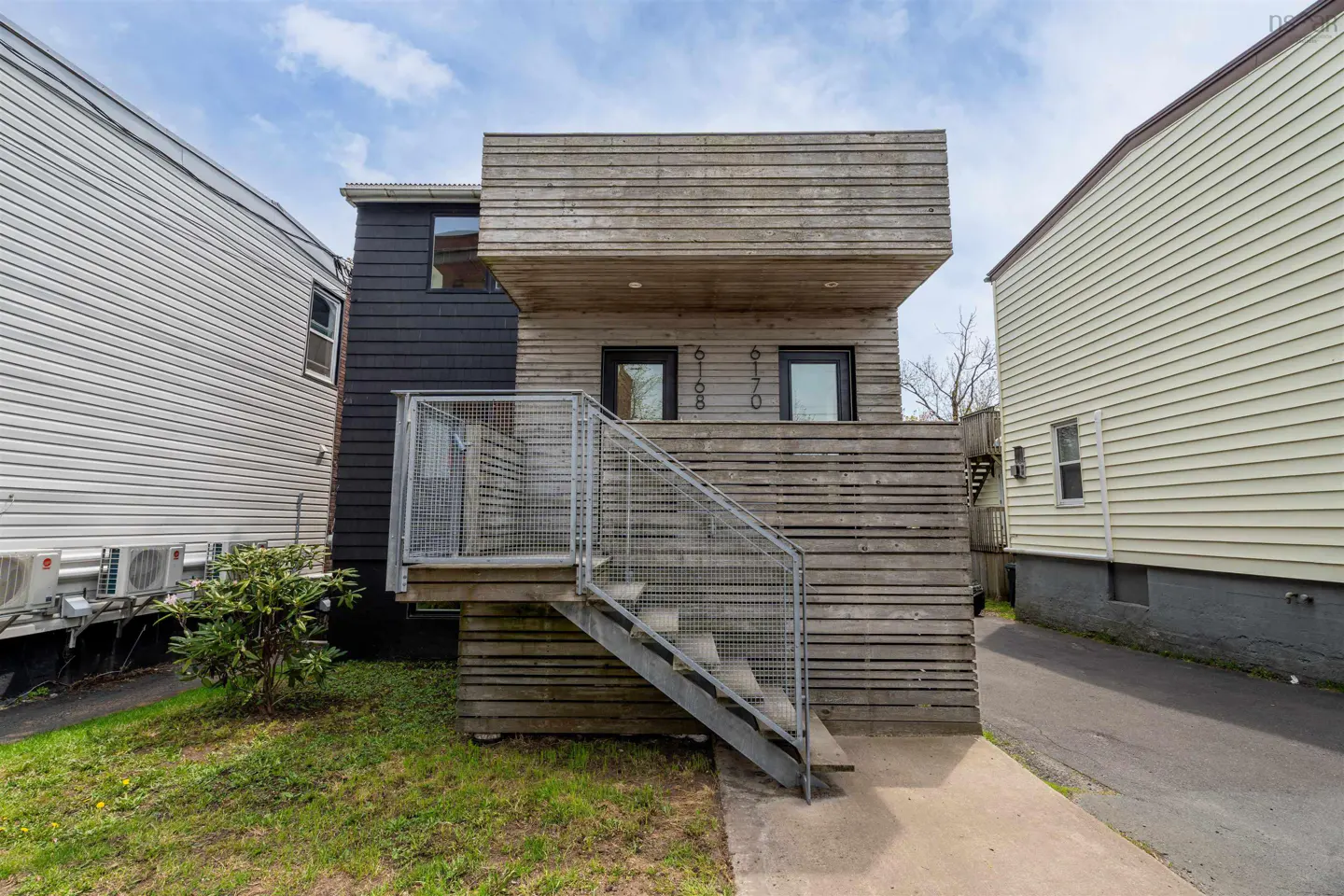 Exterior view of a modern, wood-clad duplex with metal stairs leading to the entrance. The building is flanked by two houses with siding.
