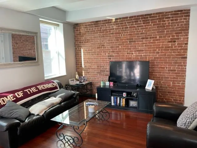 Living room with exposed brick wall, black leather sofa with "Home of the Ironmen" blanket, glass coffee table, and black TV stand.