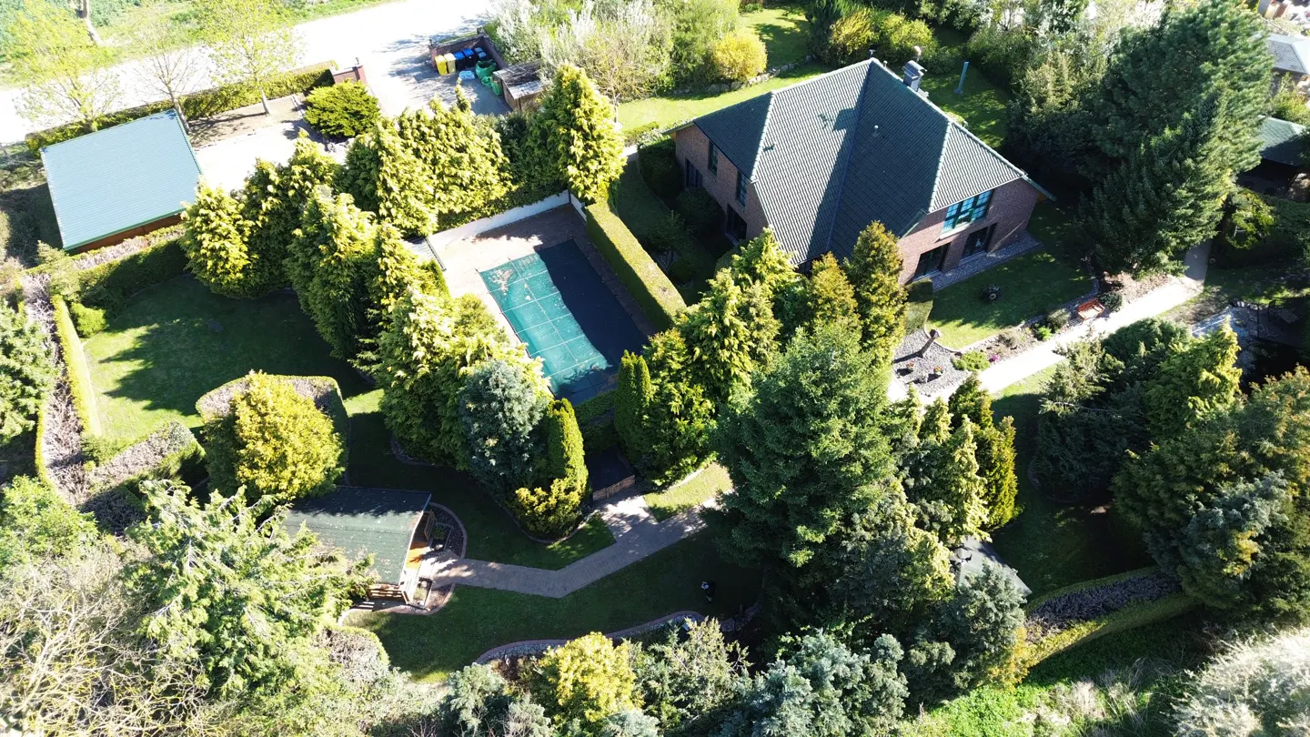 Aerial view of a brick house with a dark green roof, surrounded by lush green trees, a pool with a cover, and a small wooden structure.