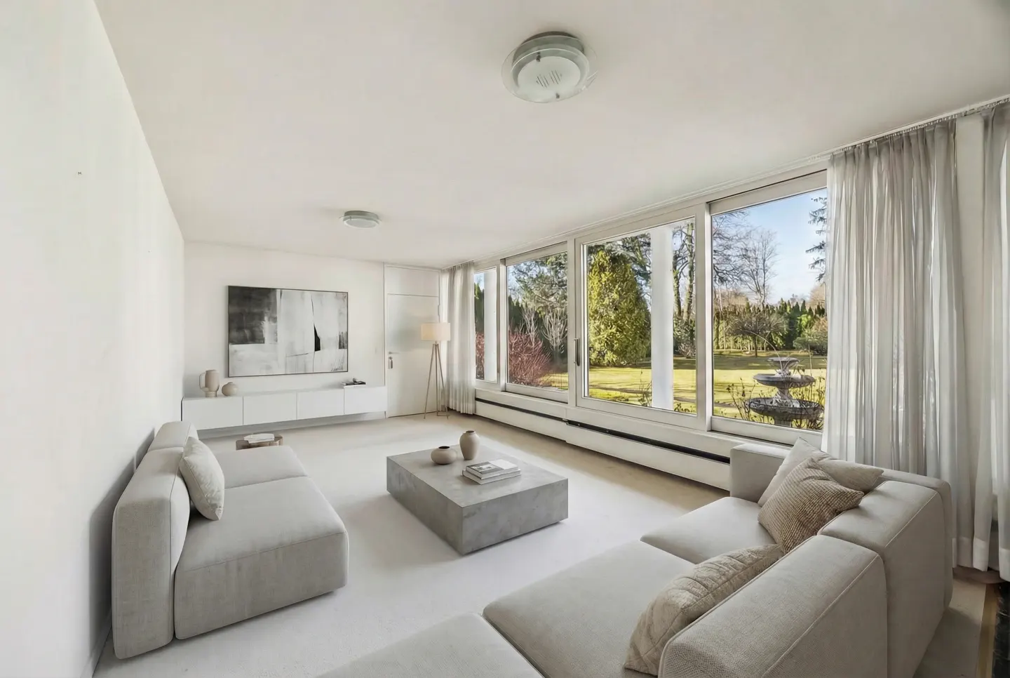 Bright living room with white walls, sofas, and a concrete coffee table. Large windows overlook a green lawn with a fountain.