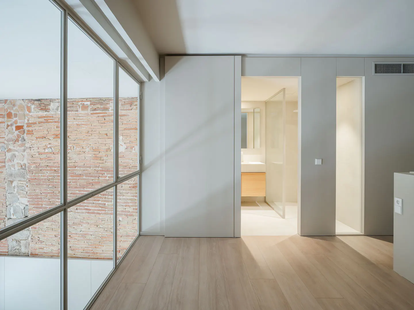 Minimalist interior with light wood floors, white walls, and large windows showing a brick building. Open doorways lead to a bathroom and another room.