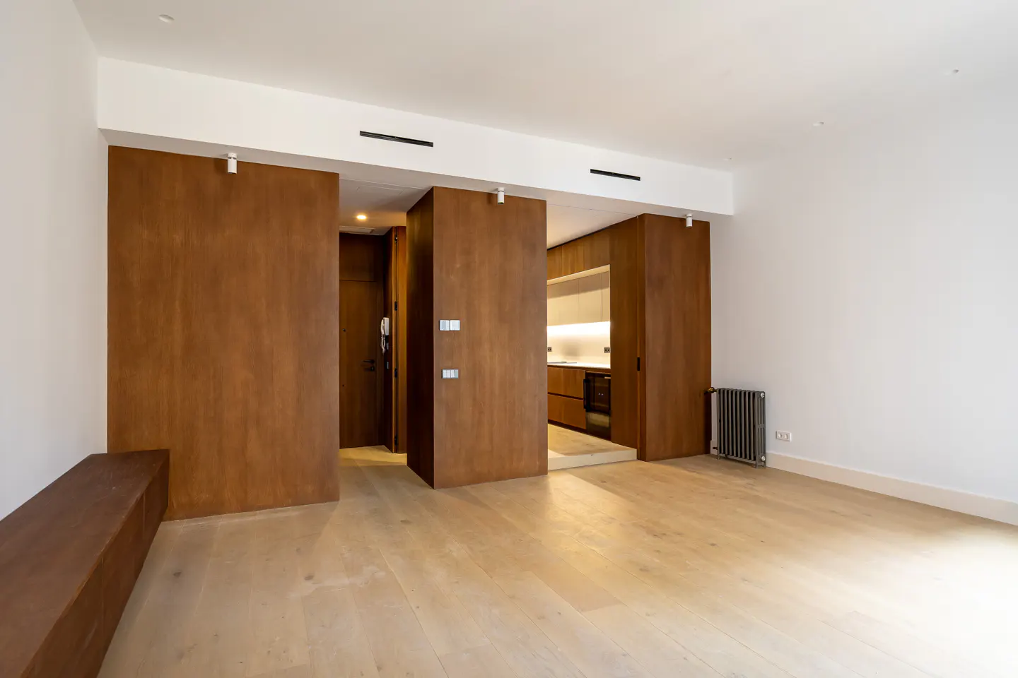 Bright, empty apartment with light wood floors, white walls, and brown wood sliding doors to the kitchen and entry. A brown bench sits against the wall.