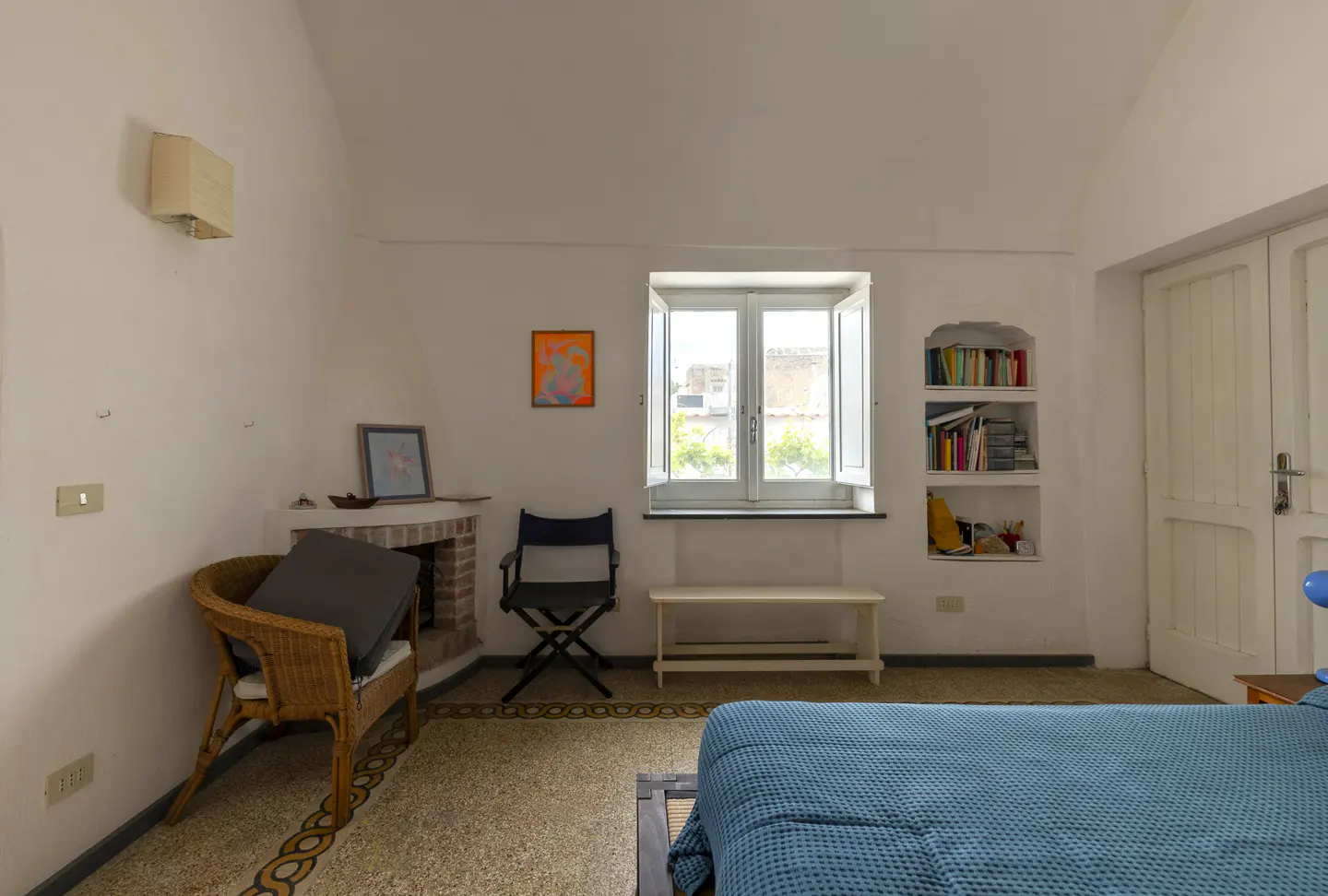 Bedroom with blue bedspread, wicker chair, fireplace, and window with white shutters. Bookshelves are built into the wall.