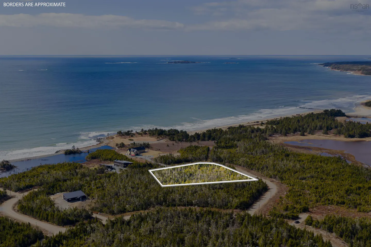 Aerial view of a wooded lot outlined in white, near the ocean. Houses are visible in the distance.