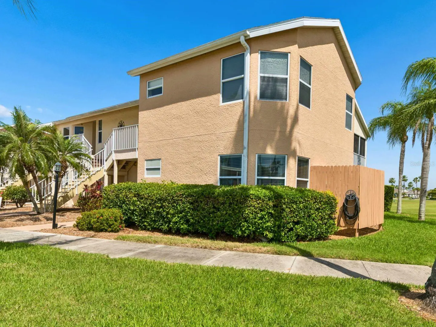 Two-story tan condo building with white trim, stairs to the second floor, green bushes, and palm trees under a blue sky.
