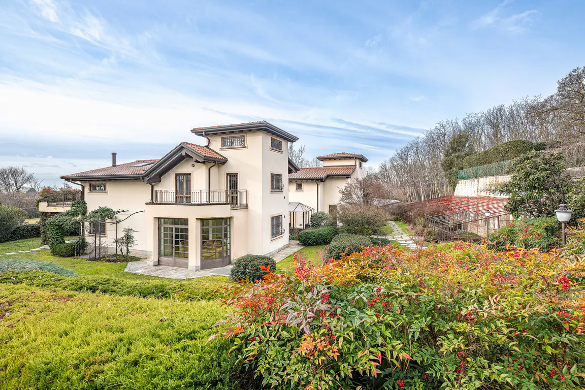 Exterior view of a multi-story cream-colored house with a red tile roof, surrounded by green lawns and colorful bushes.