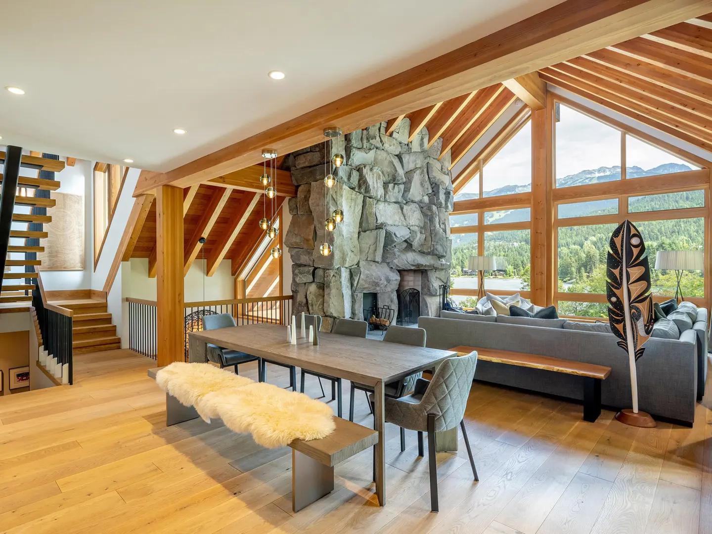 A bright, open-concept living and dining area with wood floors, exposed beams, and a stone fireplace. A long table with chairs and a bench sits in the center. Large windows offer a view of mountains.