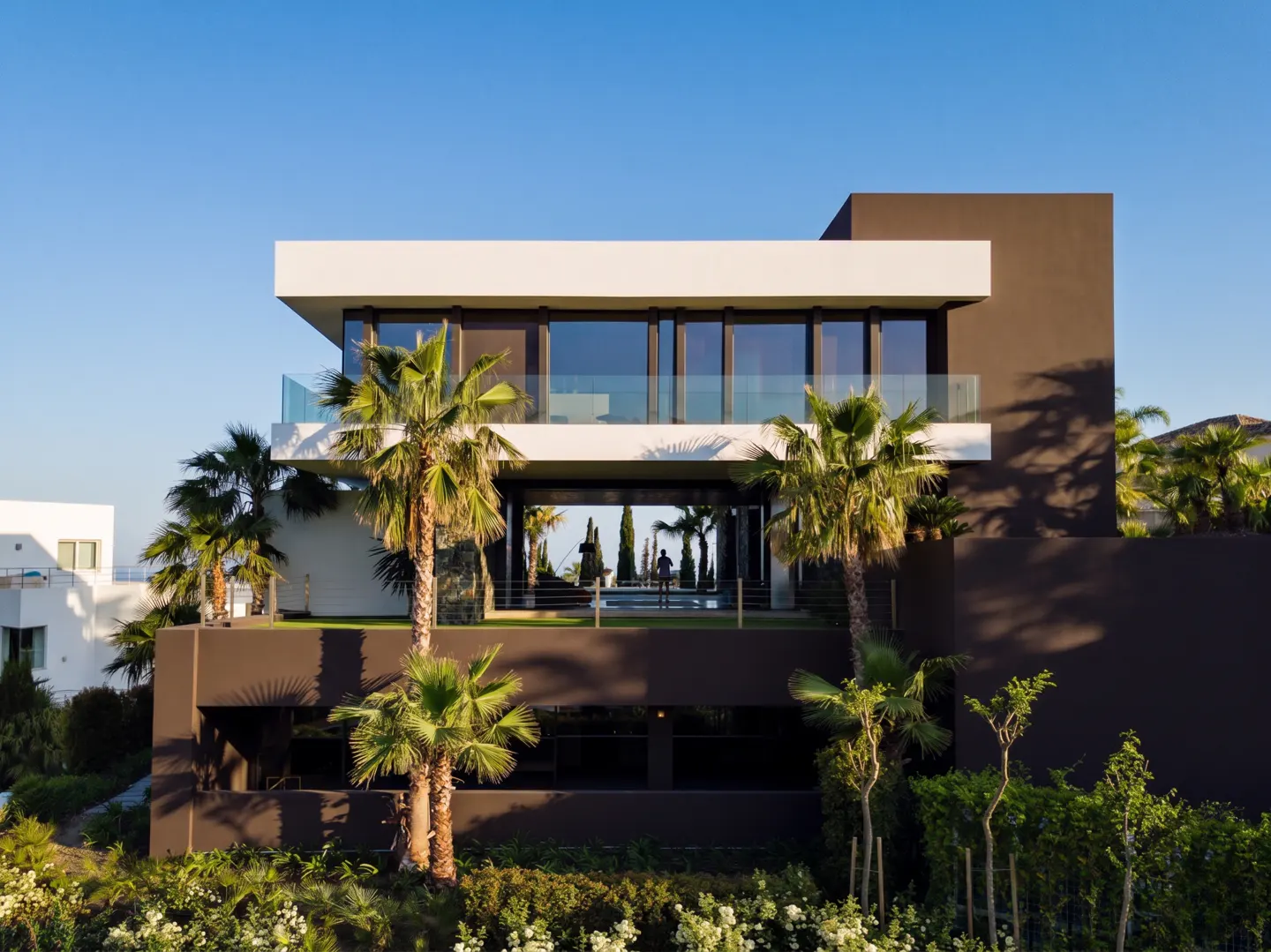 Modern two-story house with a flat roof, dark brown walls, and large windows. Palm trees and greenery surround the property.