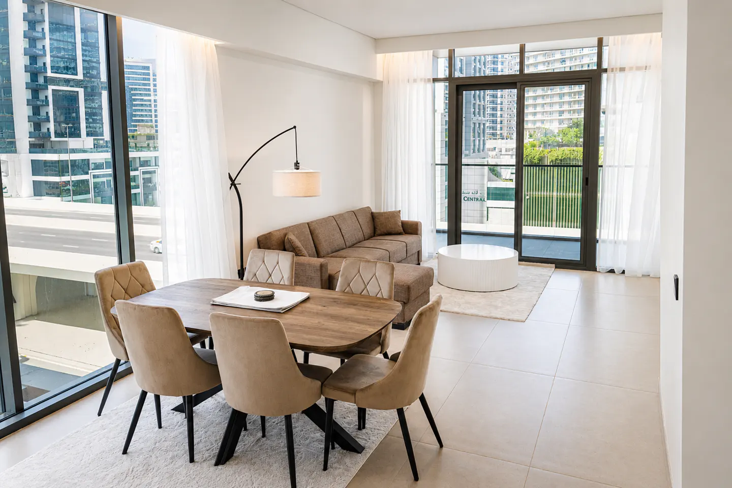 Bright apartment interior with a wooden dining table, beige chairs, brown sofa, and a white round coffee table. Large windows show a city view.