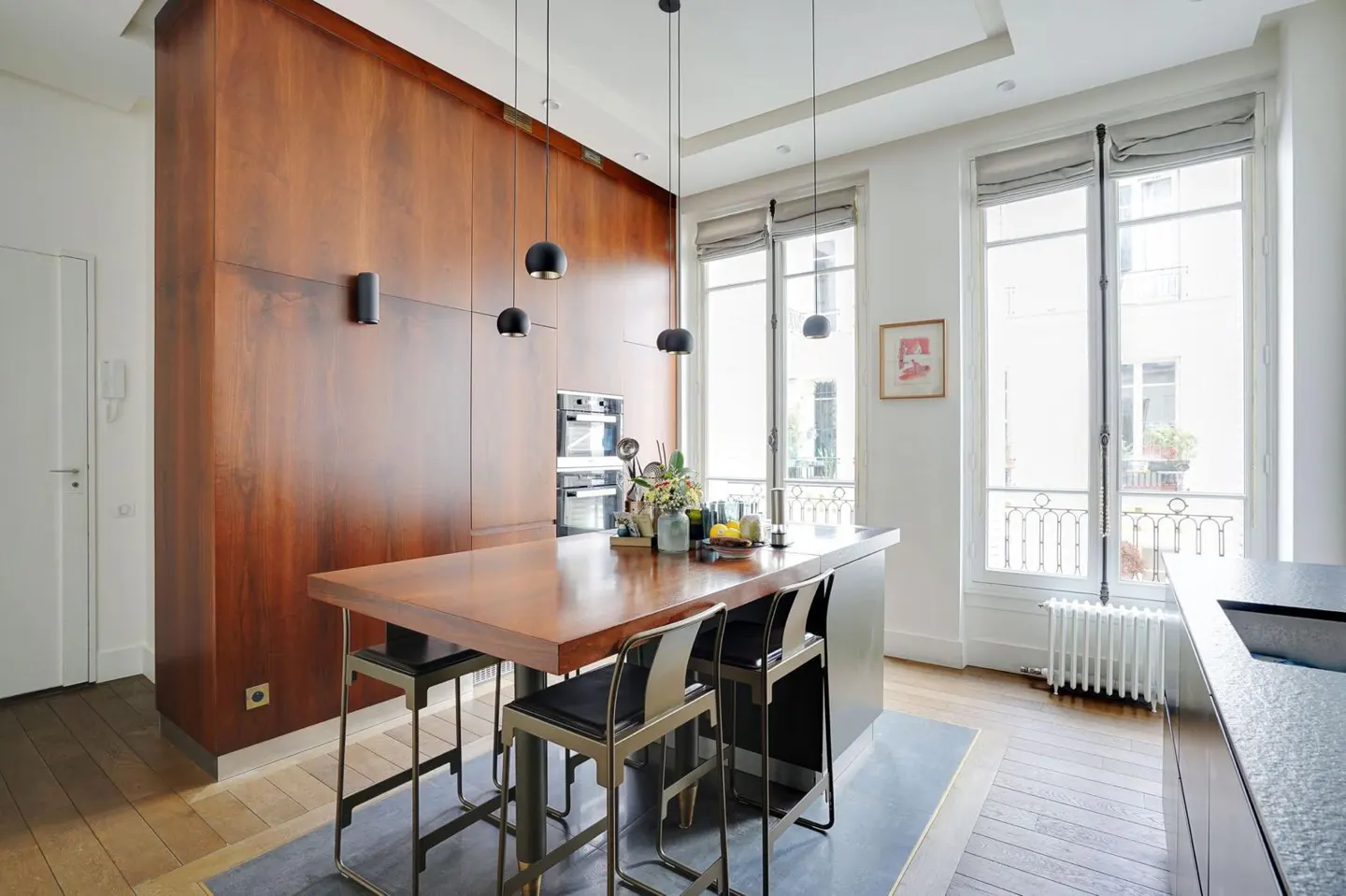 A modern kitchen with a wood-paneled wall, island with seating, and large windows. Black pendant lights hang above the island.