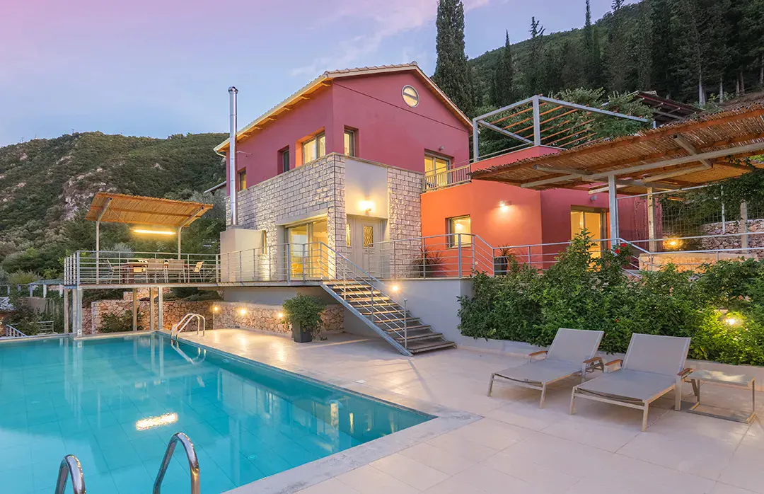 A pink and stone villa with a swimming pool, lounge chairs, and a wooden pergola at dusk against a mountain backdrop.