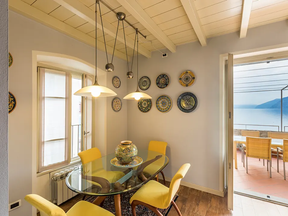Dining room with yellow chairs, glass table, and decorative plates on the wall. Balcony with lake view in the background.