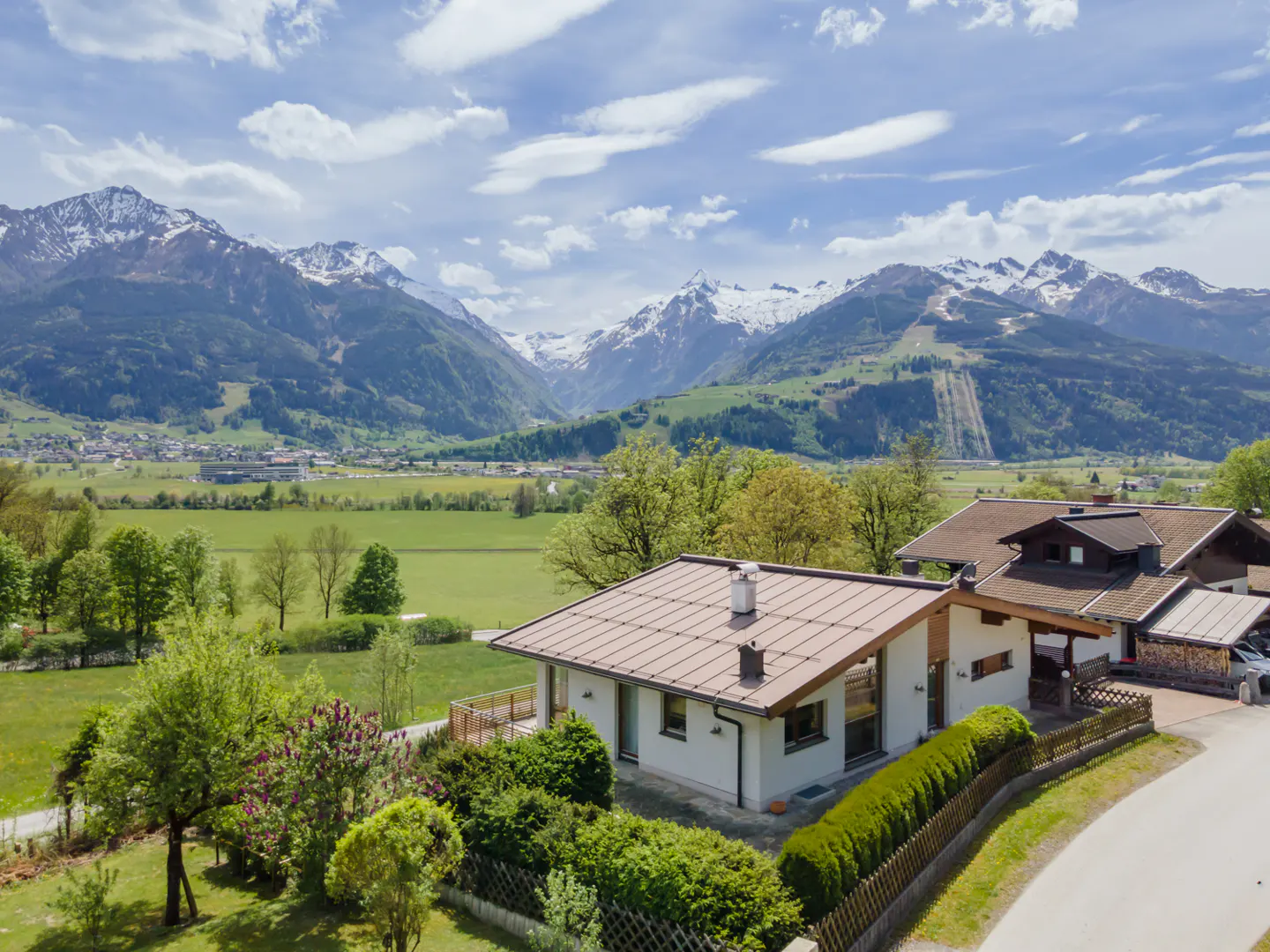 A white house with a brown roof sits in front of green fields and snow-capped mountains under a blue sky.