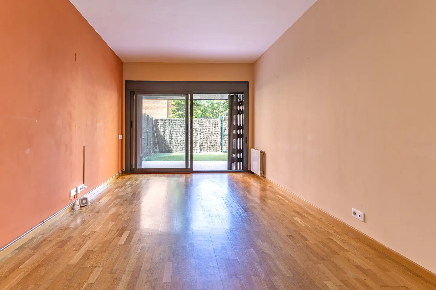 Empty room with hardwood floors, orange and beige walls, and sliding glass doors to a fenced yard.