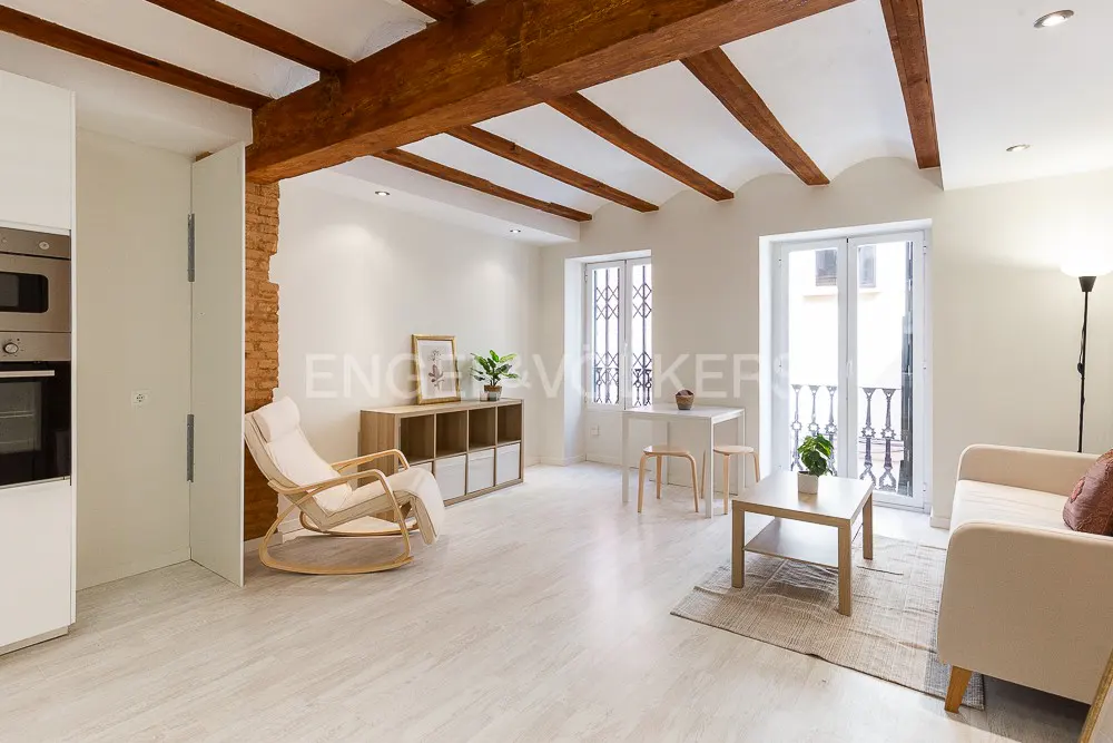 Bright living room with white walls, wood beams, and light wood furniture. A rocking chair sits near a brick accent wall. Balcony doors let in natural light.