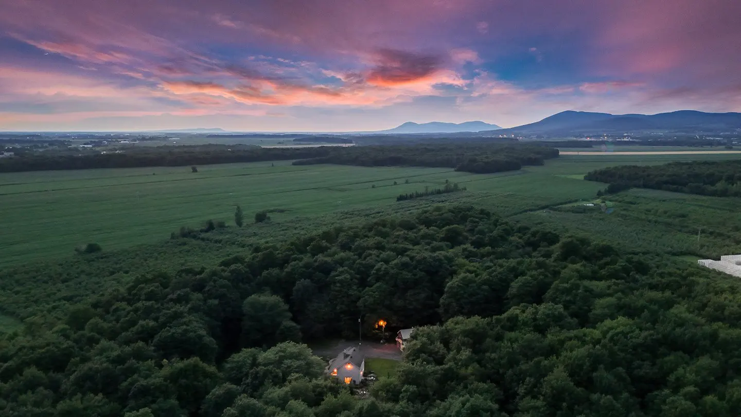 Aerial view of a house nestled in a forest, with green fields and mountains under a colorful sunset sky.