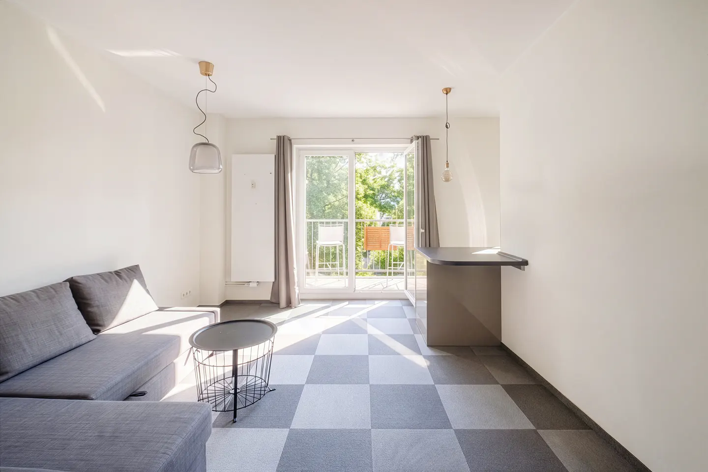 Bright, modern apartment interior with gray sofa, black table, and checkered floor. Balcony doors open to greenery.